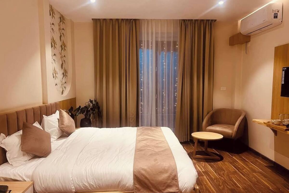 Una habitación de hotel con una cama grande, una mesita redonda pequeña, una silla y un ventanal con cortinas.