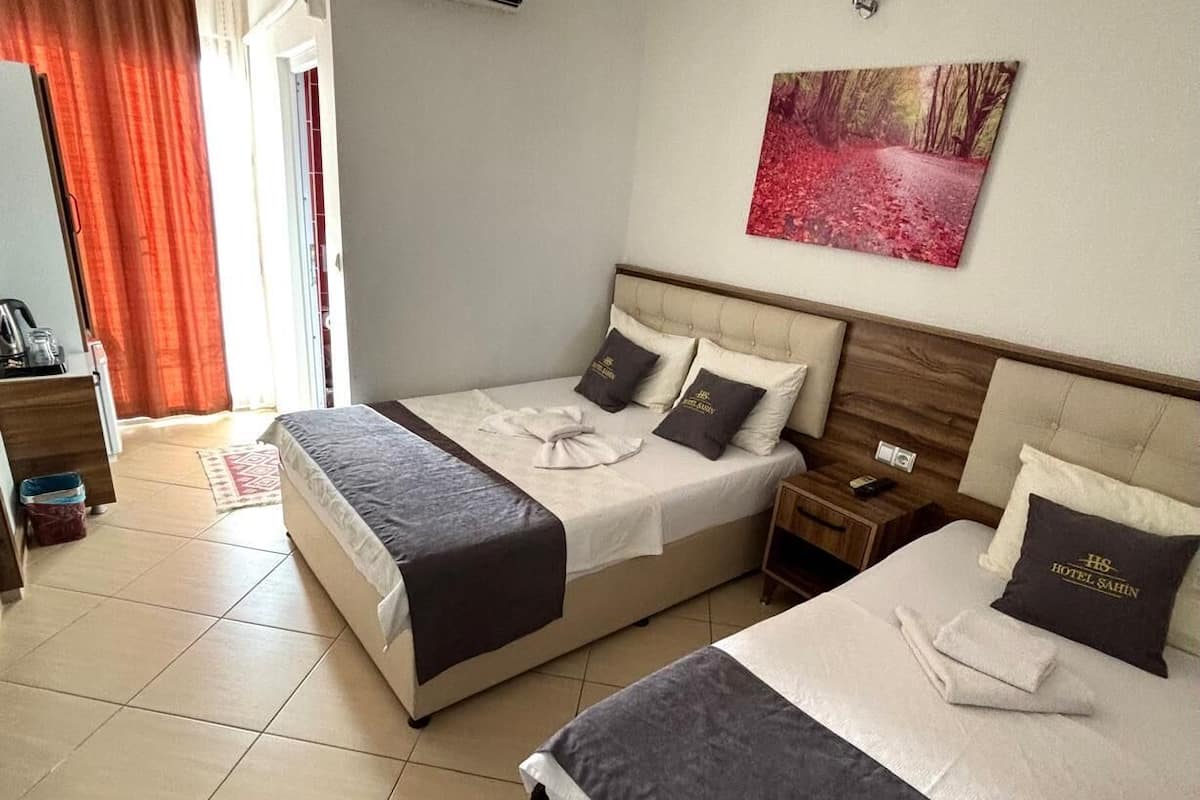 A hotel room with two beds, a painting, a bedside table, a nightstand, a window with curtains, and an air conditioning unit.