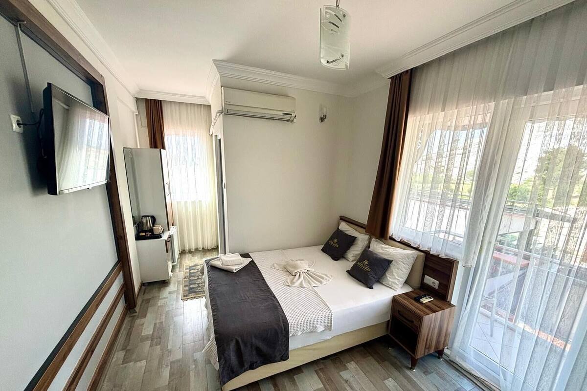 A hotel room with a bed, a TV, an air conditioning unit, a small table with a kettle, and a window with curtains.