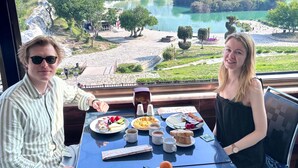 Una mesa de comedor con comida y bebidas, una vista panorámica de un lago y colinas, y una persona sentada a la mesa.