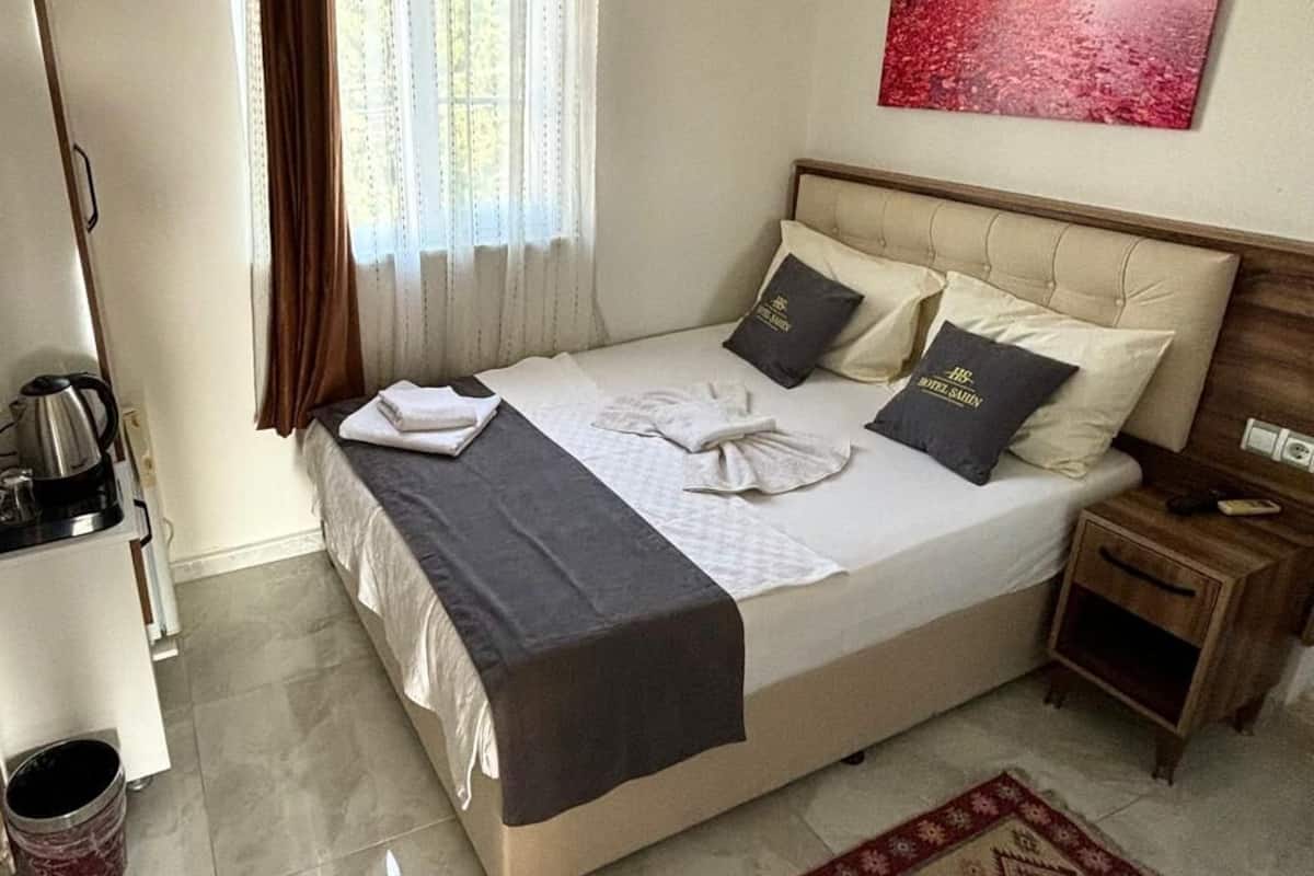 A hotel room with a bed, a nightstand, a coffee maker, a kettle, a rug, and a painting on the wall.