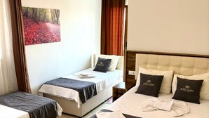 Habitación de hotel con dos camas individuales, una cama más grande y un cabecero con un logo. Hay un cuadro en la pared, una ventana con cortinas y un equipo de aire acondicionado.