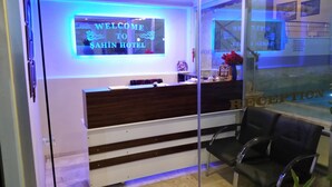 Un mostrador de recepción con un cartel que dice "WELCOME TO SAHIN HOTEL" y un área de espera con sillas y mostrador.