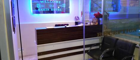 A reception desk with a sign reading "WELCOME TO SAHIN HOTEL" and a reception area with chairs and a counter.