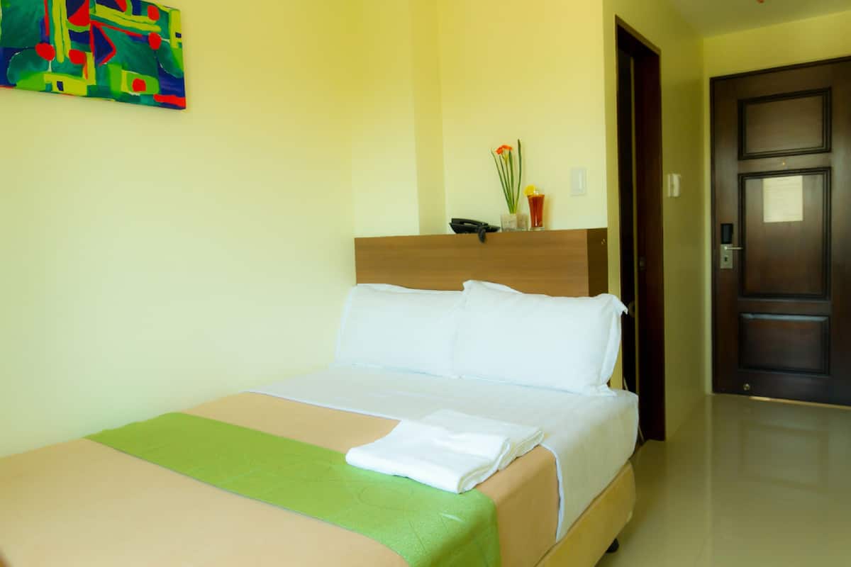A hotel room with a bed, a green throw blanket, a white pillow, and a vase with flowers on the nightstand.