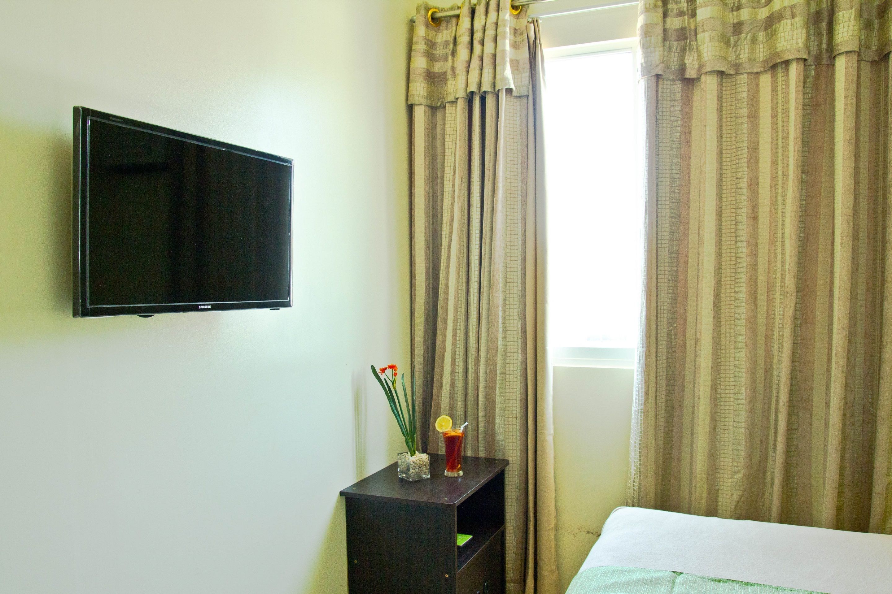 A hotel room with a flat-screen TV mounted on the wall, a small table with a vase of flowers and a drink, and a bed with a white sheet.