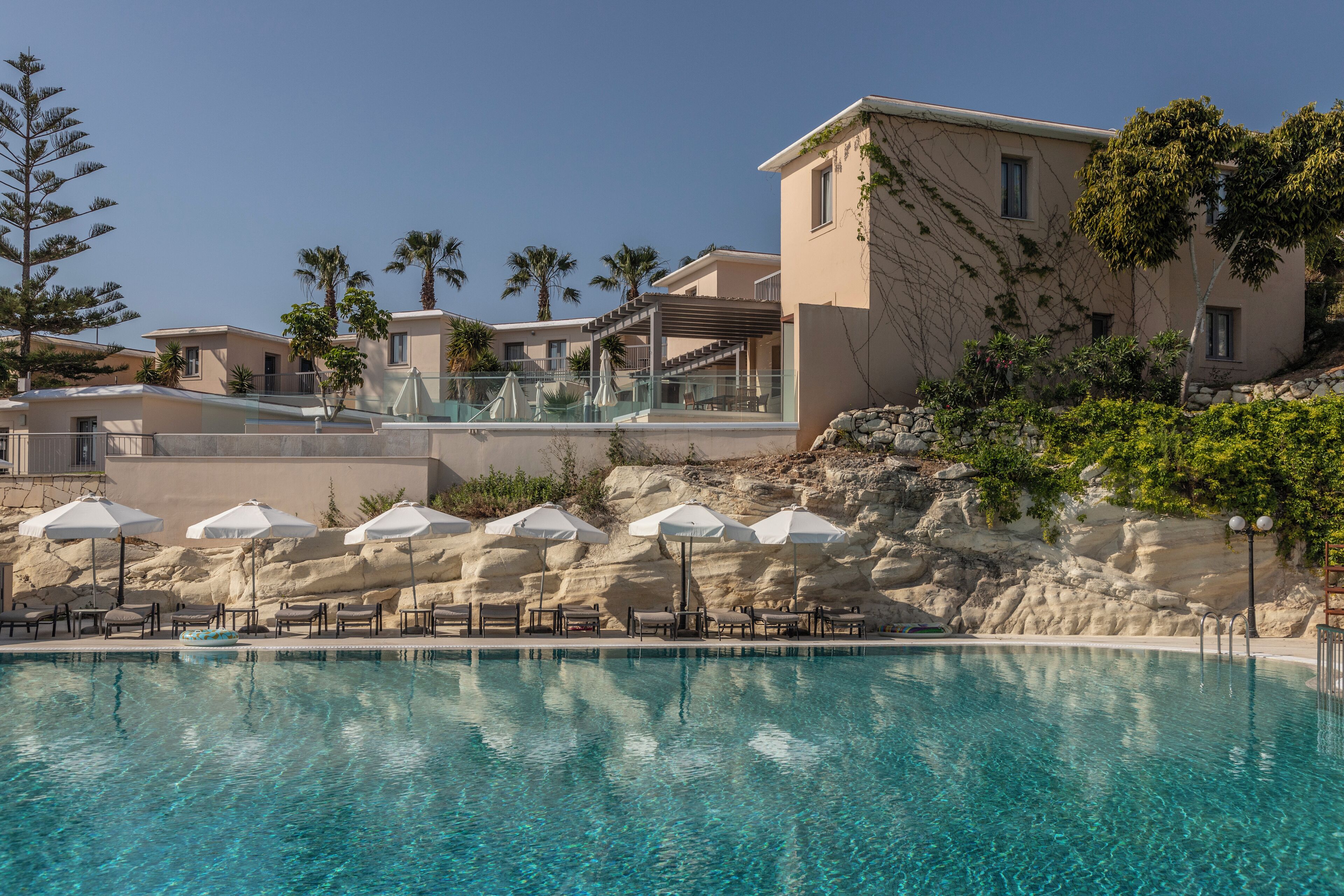 Zona de piscina con sillas de descanso y sombrillas, rodeada de edificios de estilo mediterráneo.