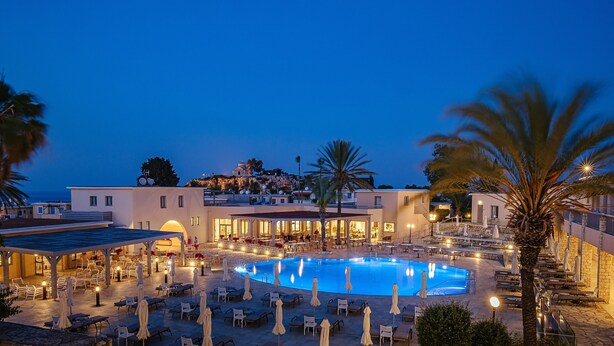 A resort pool area with sun loungers and umbrellas, a building with a pool, and a view of a hill with a church. - Louis St Elias Resort & Waterpark (Paralimni)