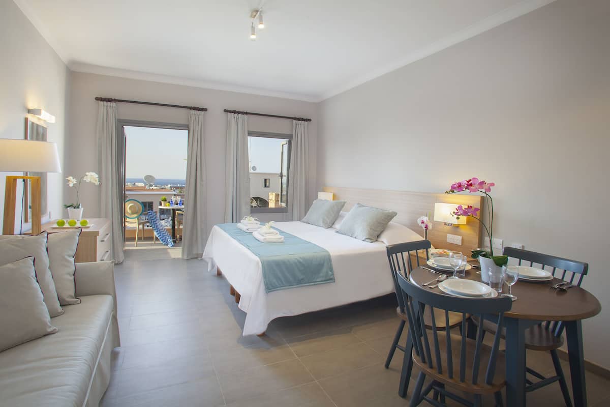 Quarto de hotel com cama, área de estar com mesa e cadeiras, varanda com vista e pequena mesa com abajur e flores.