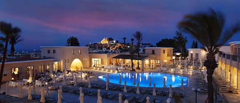 A resort pool area with lounge chairs and umbrellas, surrounded by buildings and palm trees.