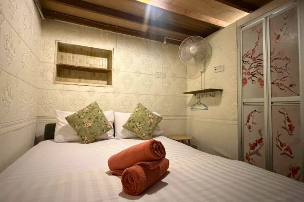 Un dormitorio con cama, almohadas, una toalla enrollada, un ventilador, una estantería y un armario.