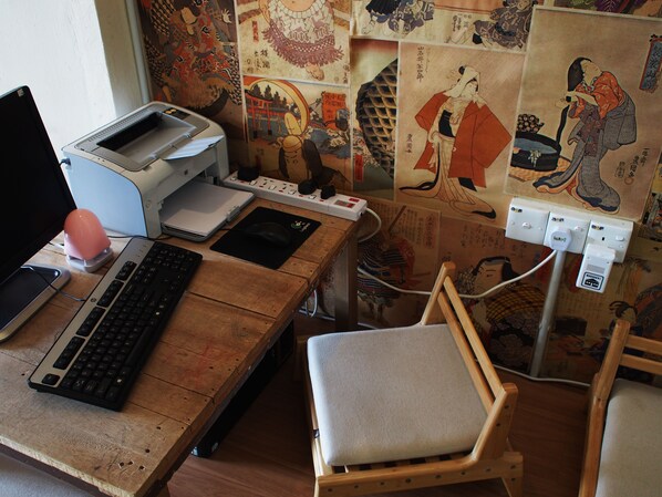 A wooden desk with a laptop, keyboard, mouse, and printer. There is a chair with a cushion. The wall behind the desk features traditional Japanese artwork. - Dou Houz - Hostel (George Town)
