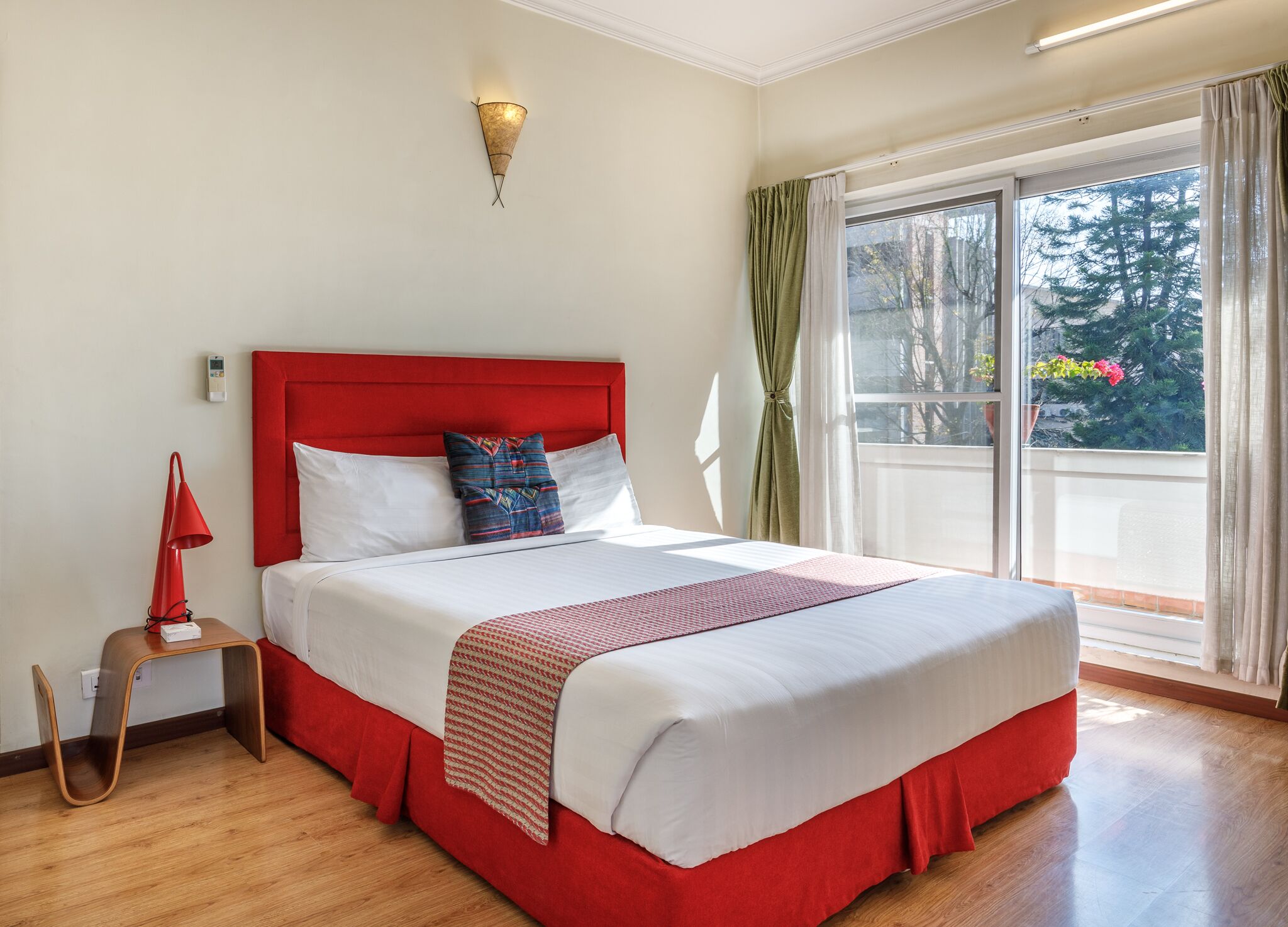A hotel room with a large bed, a red headboard, a bedside table with a lamp, and a view of the outdoors through a sliding glass door.