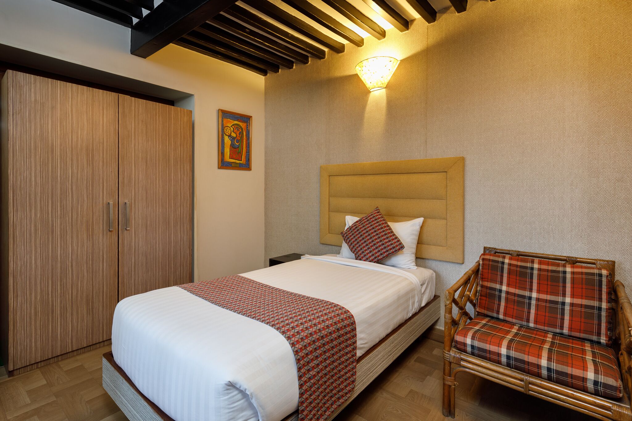 A neatly made bed with a patterned bedspread, a wooden wardrobe, a wicker chair, and a wall-mounted light fixture.