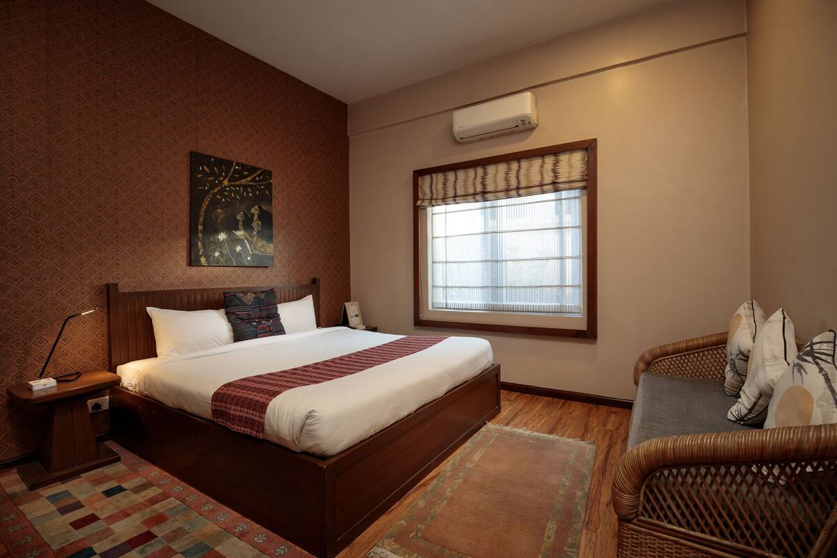 A bedroom with a large bed, a nightstand, a wicker chair, a window with blinds, and a wall-mounted air conditioner.