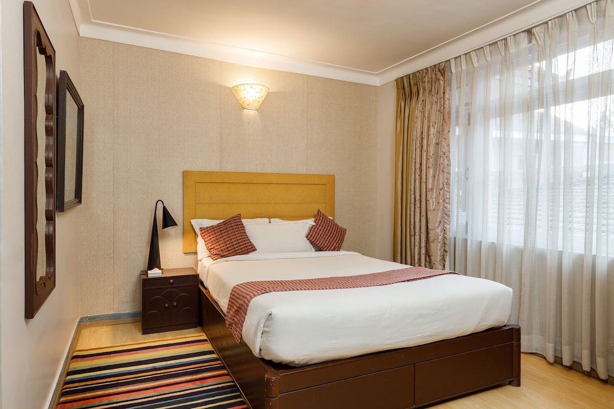 A hotel room with a large bed, a nightstand, a lamp, a striped rug, and a window with curtains.