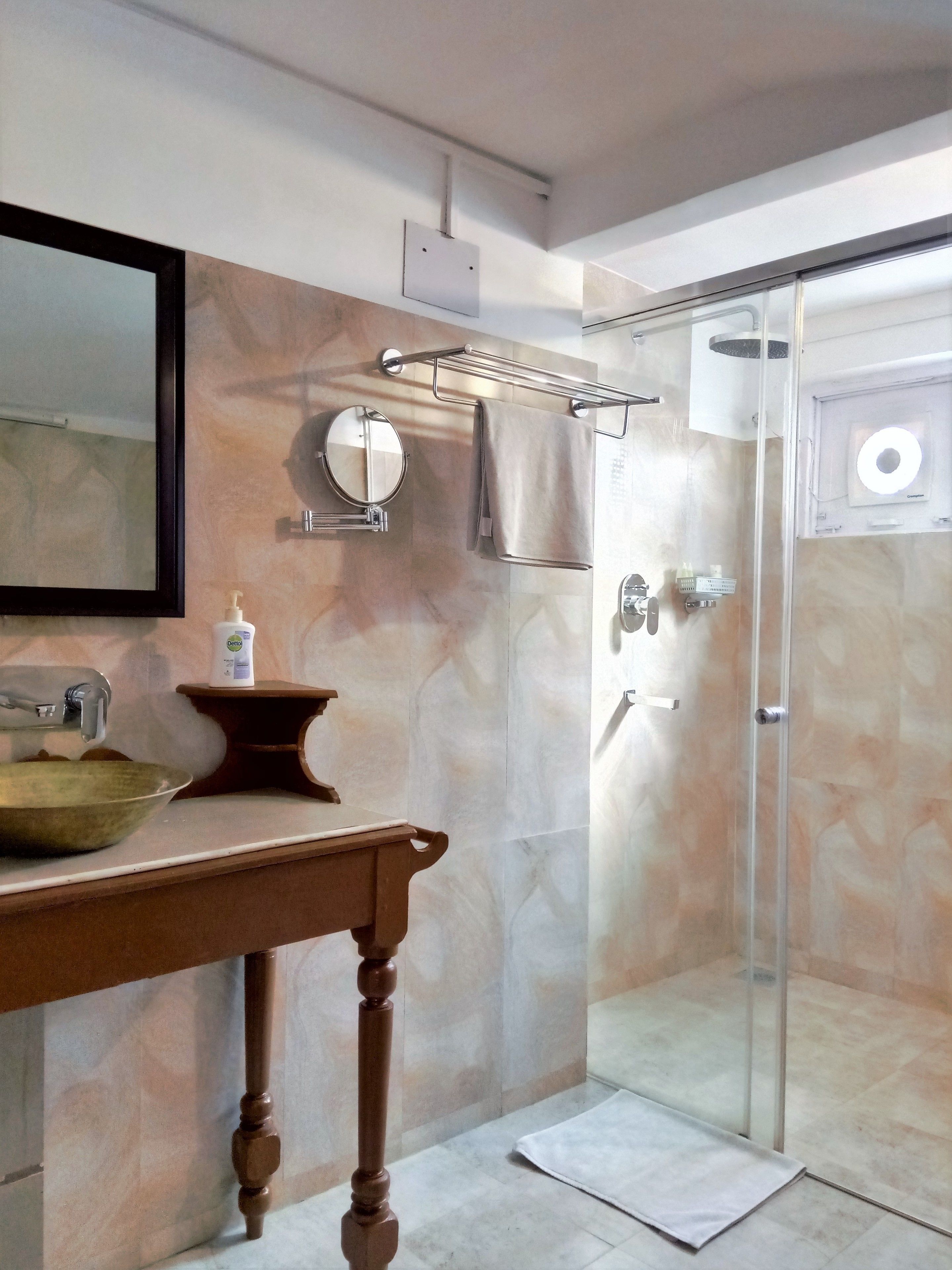 Une salle de bain avec un mur en marbre, un meuble-lavabo en bois, un miroir rond, un porte-serviettes, une cabine de douche en verre et une douchette à main.