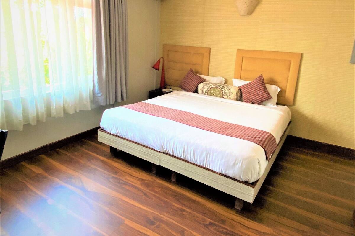 A hotel room with a bed, wooden flooring, a ceiling light, an air conditioning unit, and a window with curtains.