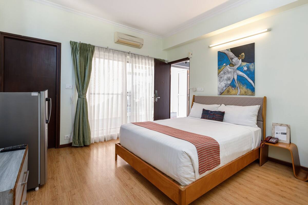 A hotel room with a wooden bed, a refrigerator, a bedside table with a phone, and a painting on the wall.