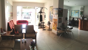 Un vestíbulo de hotel con mostrador de recepción, sillas, una pequeña mesa con un florero, una zona de gimnasio con equipo de ejercicio y un amplio ventanal con vistas a la calle.