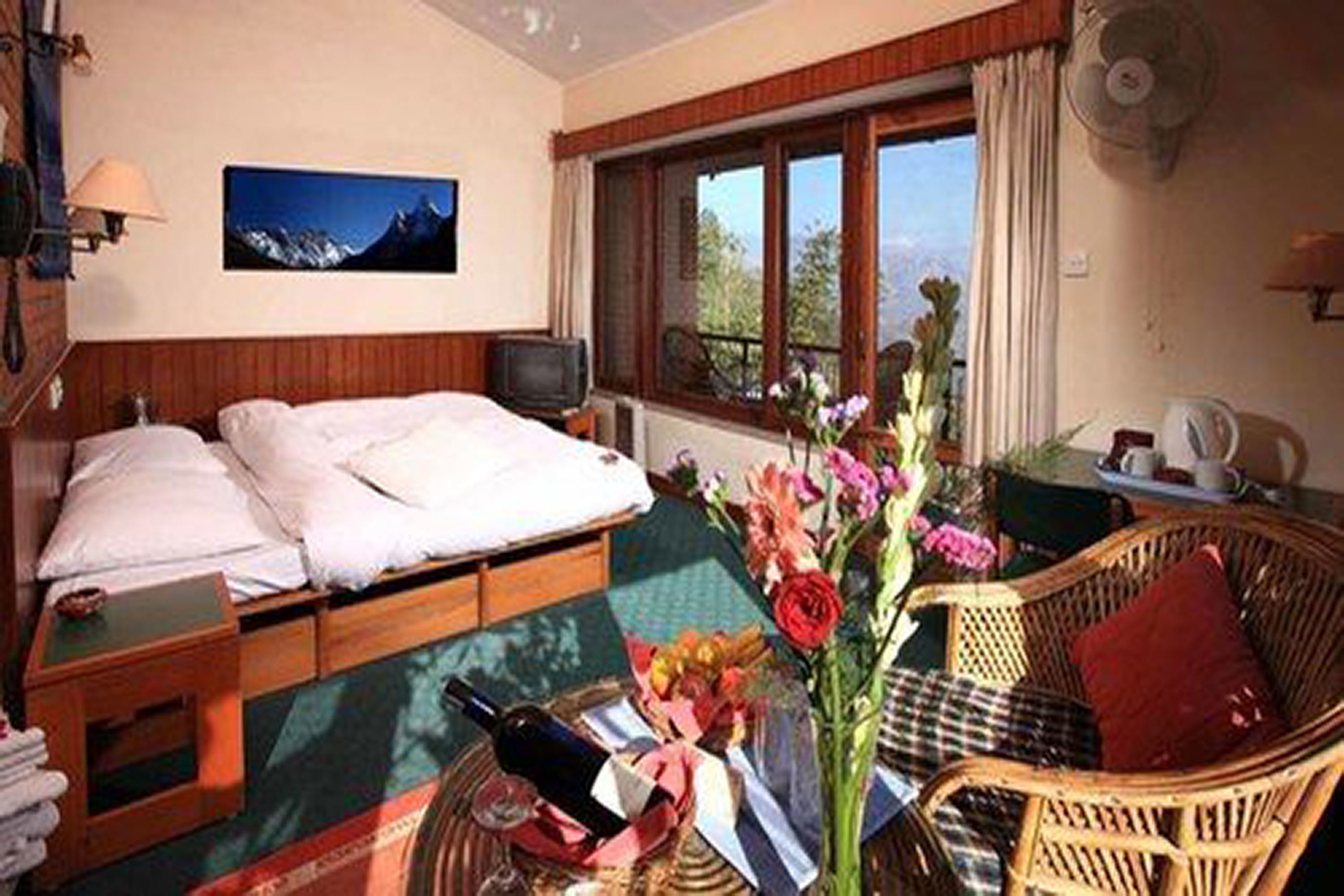 A hotel room with a bed, a television, a window with a view, a wicker chair, and a vase with flowers.