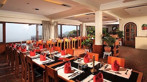 A spacious dining area with multiple tables set for service, large windows offering a view, and a potted plant in the corner.