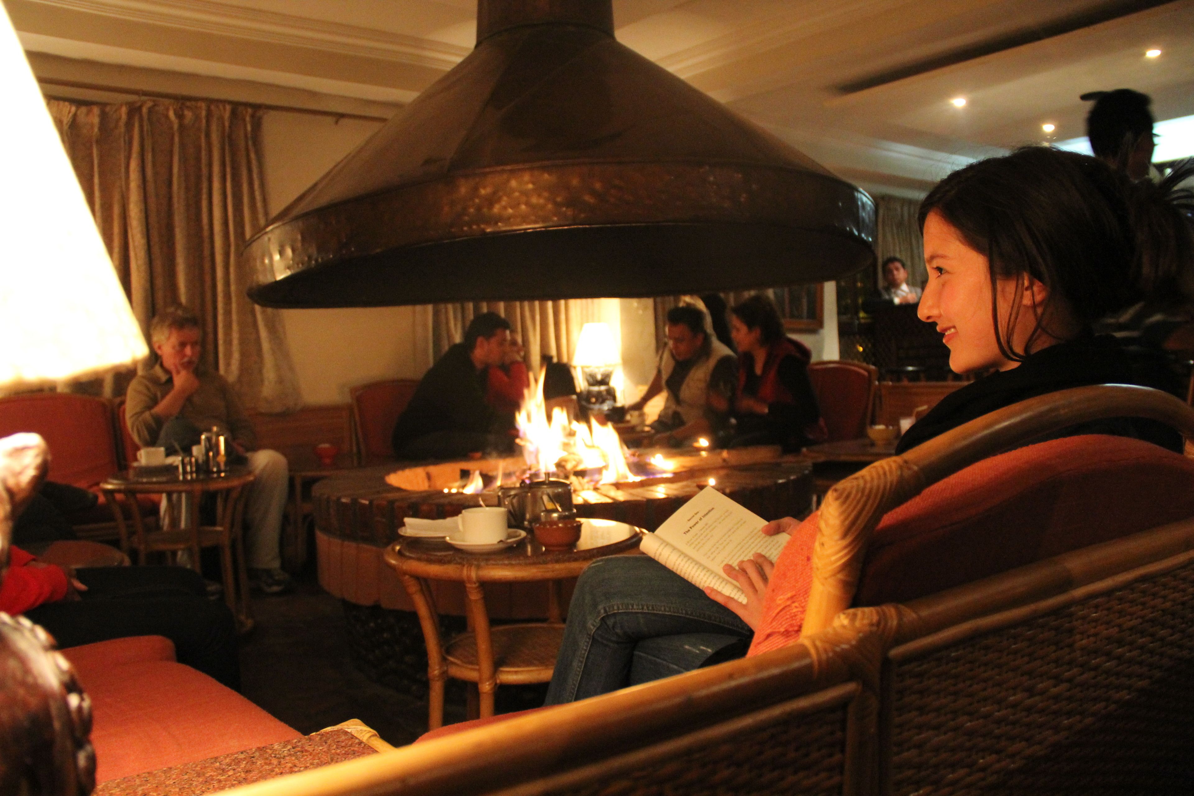 A cozy indoor setting with a central fire feature, seating arrangements, and a person reading a book.