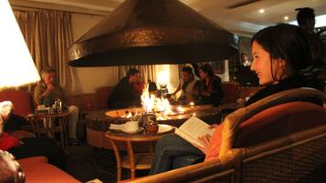 A cozy indoor setting with a central fire feature, seating arrangements, and a person reading a book.