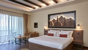 A modern hotel room with a large bed, a wooden headboard, a small table, and a chair. There is a painting on the wall and a window with curtains.