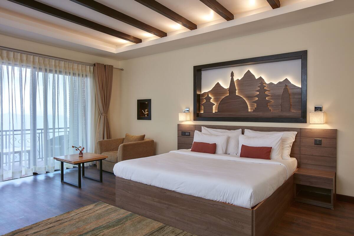 A modern hotel room with a large bed, a wooden headboard, a small table, and a chair. There is a painting on the wall and a window with curtains.