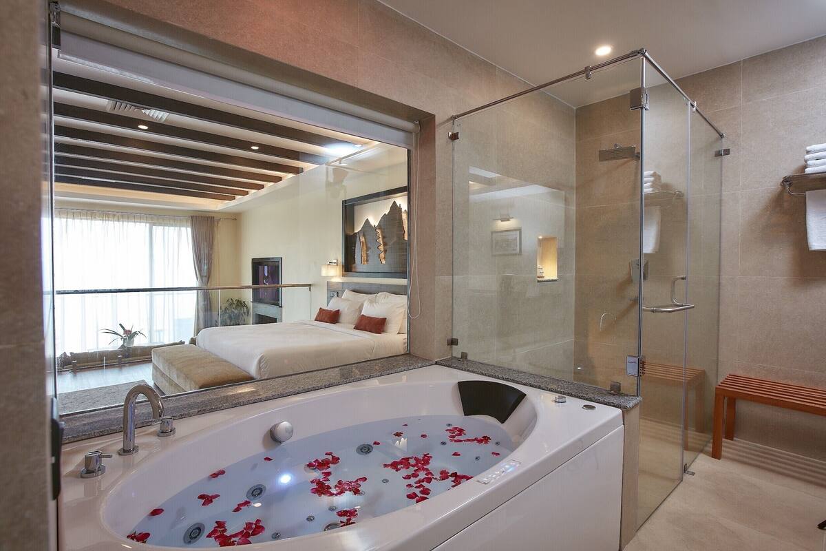 A modern bathroom with a glass-enclosed shower, a large bathtub with rose petals, a mirror, and a bed with a view of a balcony.