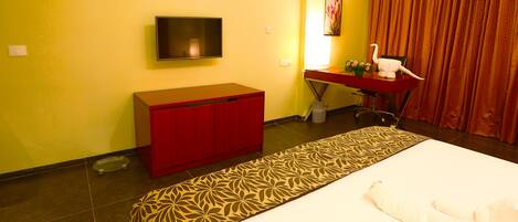 A hotel room with a bed, a red desk, a television, and a patterned rug.