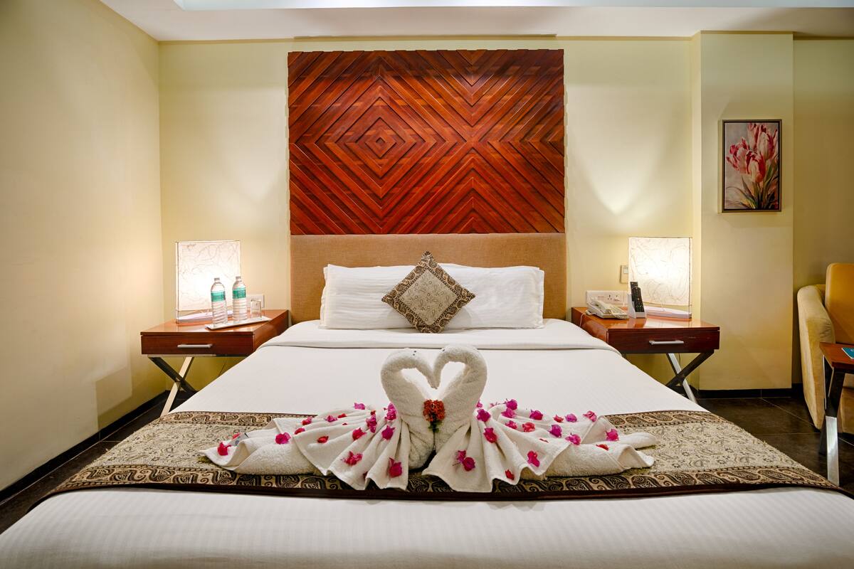 Quarto de hotel com uma cama grande, cabeceira de madeira, duas mesas de cabeceira com luminárias e um adorno floral na parede.