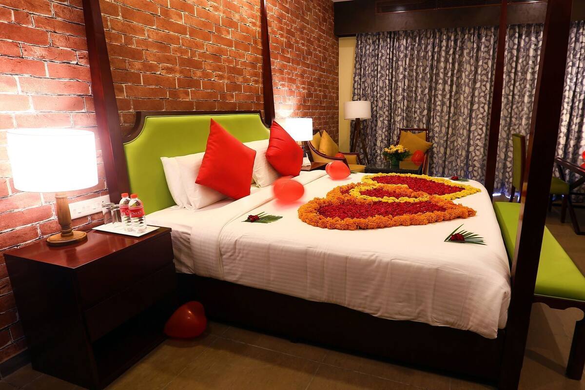 Quarto de hotel com parede de tijolos, cabeceira estofada verde, colcha branca, almofadas vermelhas, arranjo de flores em formato de coração, mesa de cabeceira com abajur e uma pequena mesa com uma almofada amarela.