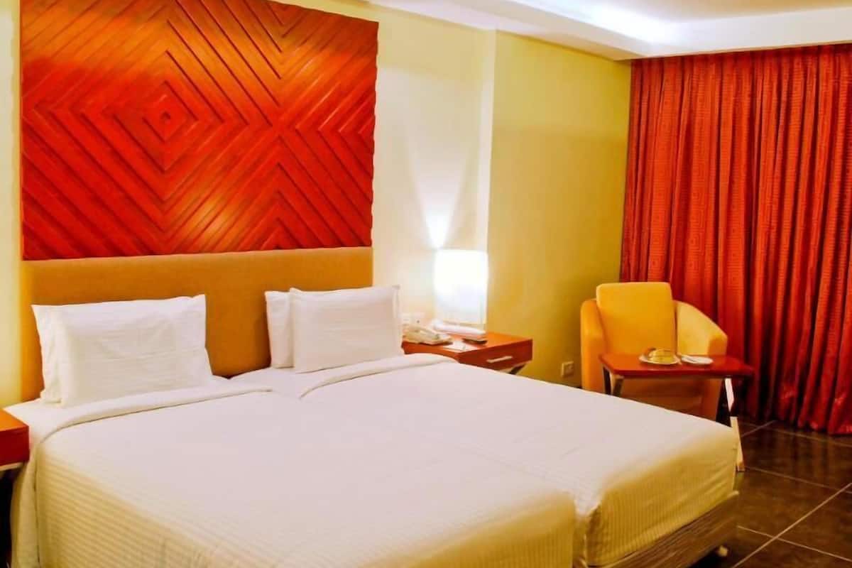 Quarto de hotel com uma cama grande, cabeceira com padrão geométrico, uma mesa pequena com uma cadeira e cortina vermelha.