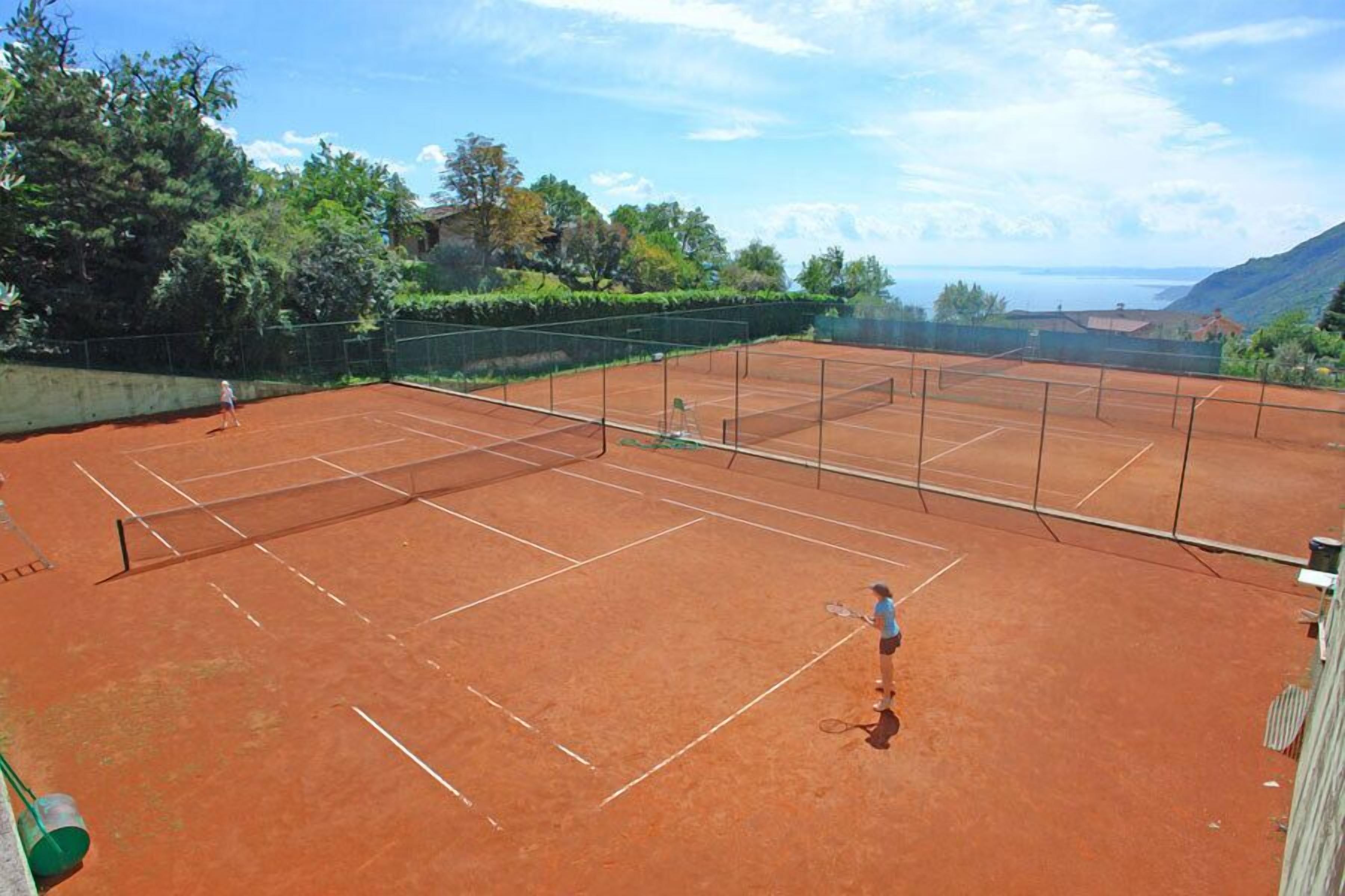 A tennis court with a net, surrounded by greenery and a view of the sea.