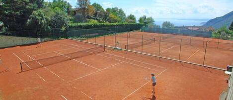 A tennis court with a net, surrounded by greenery and a view of the sea.