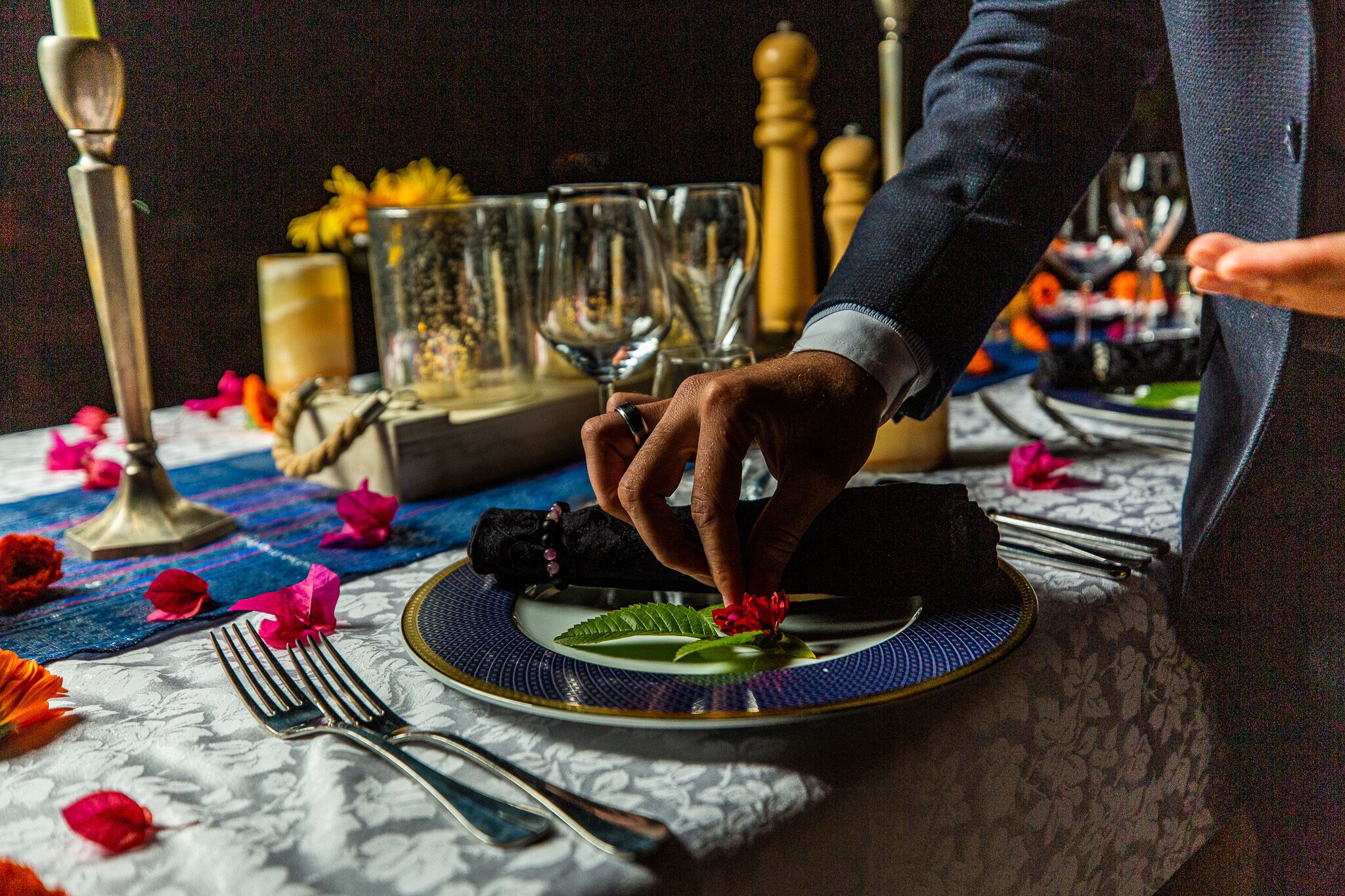 A dining table set with a plate, fork, and a hand placing a flower on the plate.