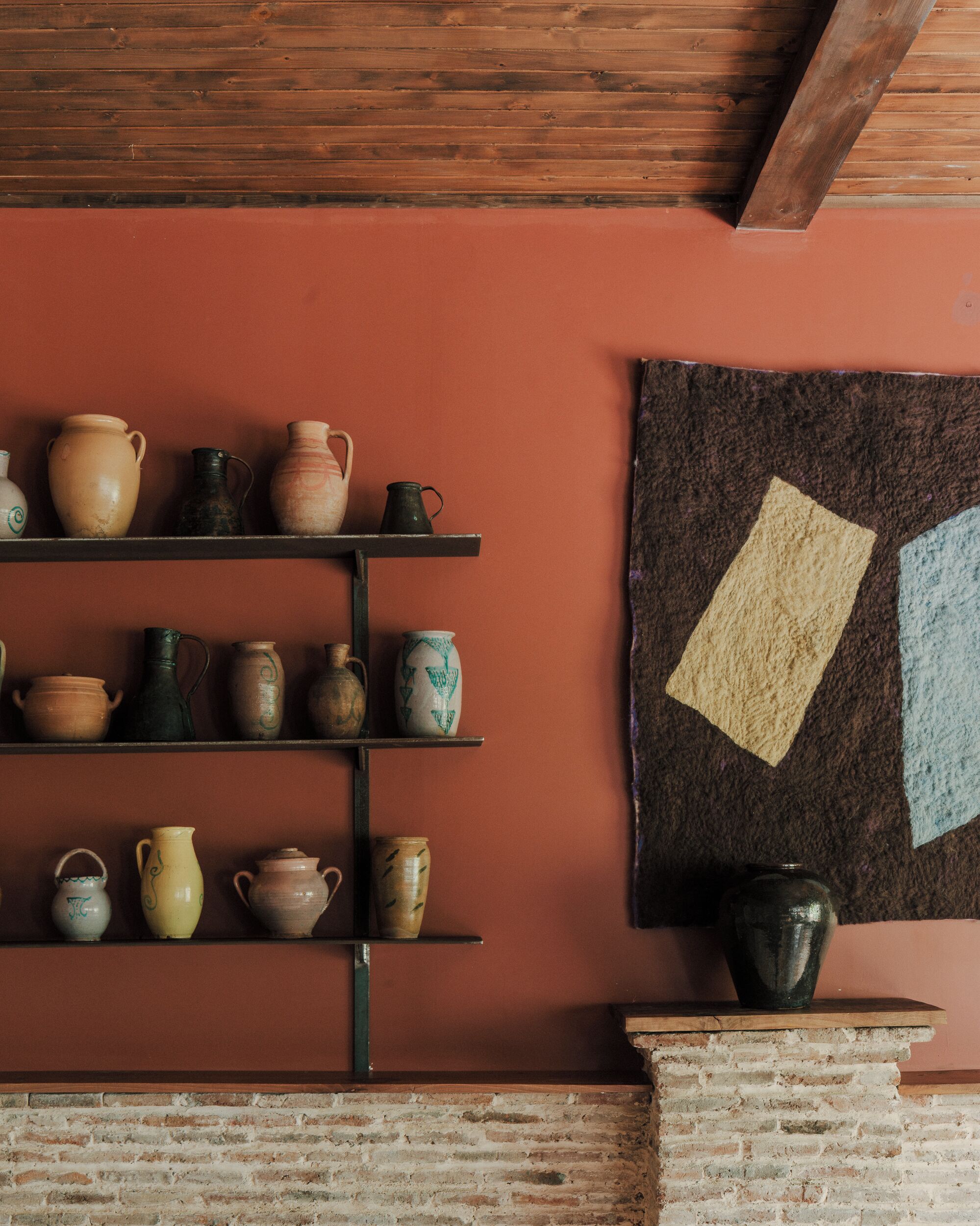 A room with a brick fireplace, shelves with pottery, and a wall with a geometric tapestry.