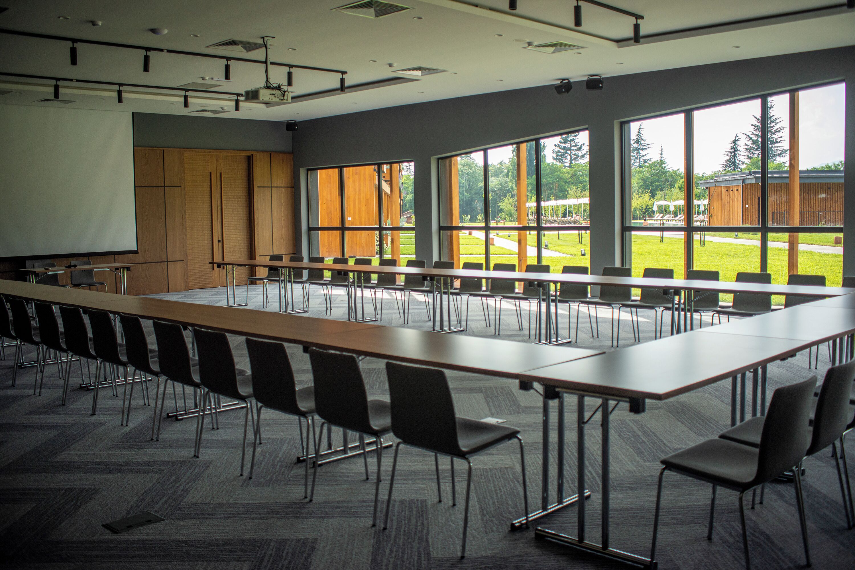 Un salón de conferencias con una larga mesa, sillas, una pantalla de proyector y ventanas amplias.