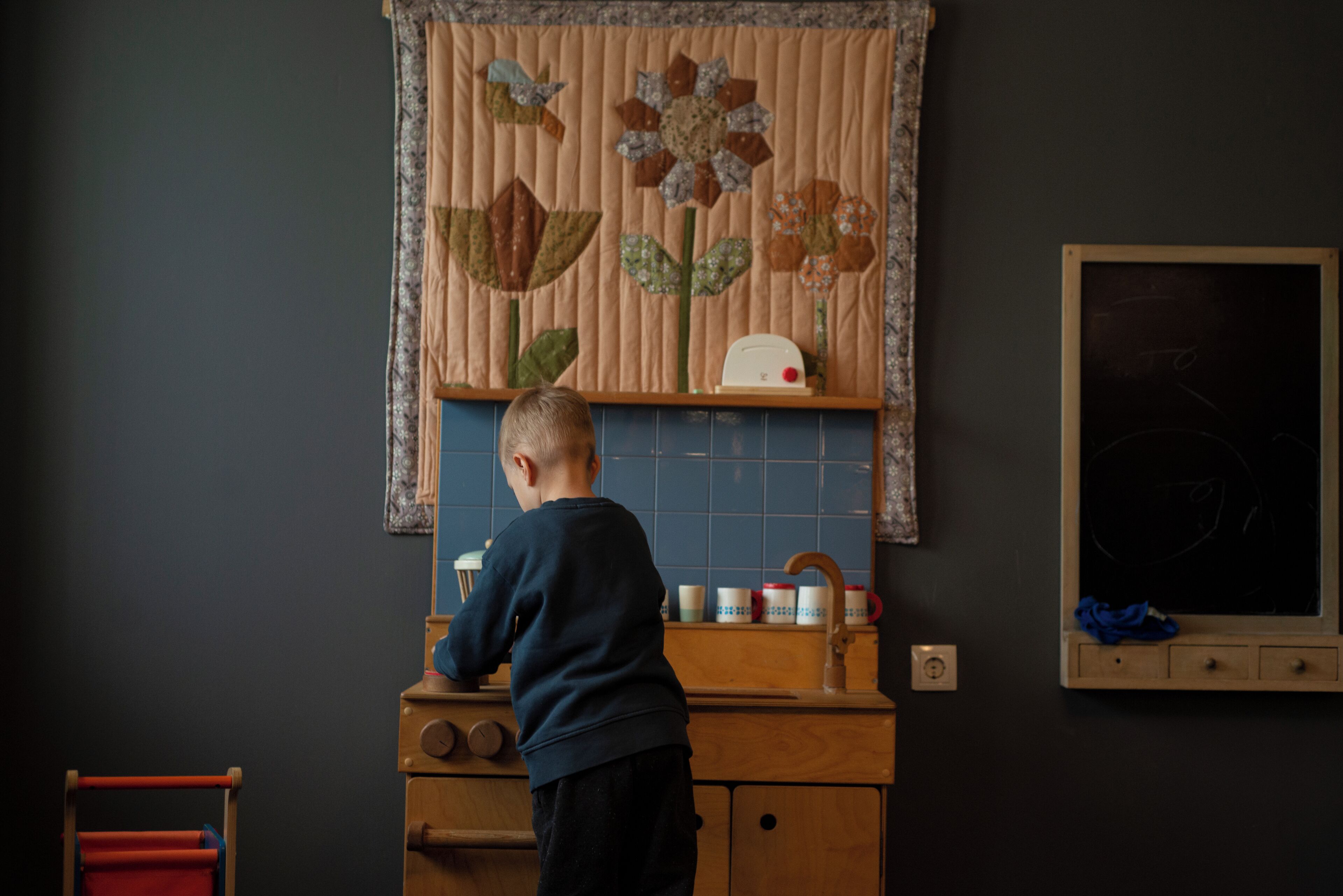 A child at a wooden kitchen counter with a sink, a window with blinds, and a wall-mounted chalkboard.
