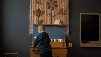 Uma criança em uma bancada de cozinha de madeira, com uma pia, uma janela com persianas e um quadro negro fixado na parede.