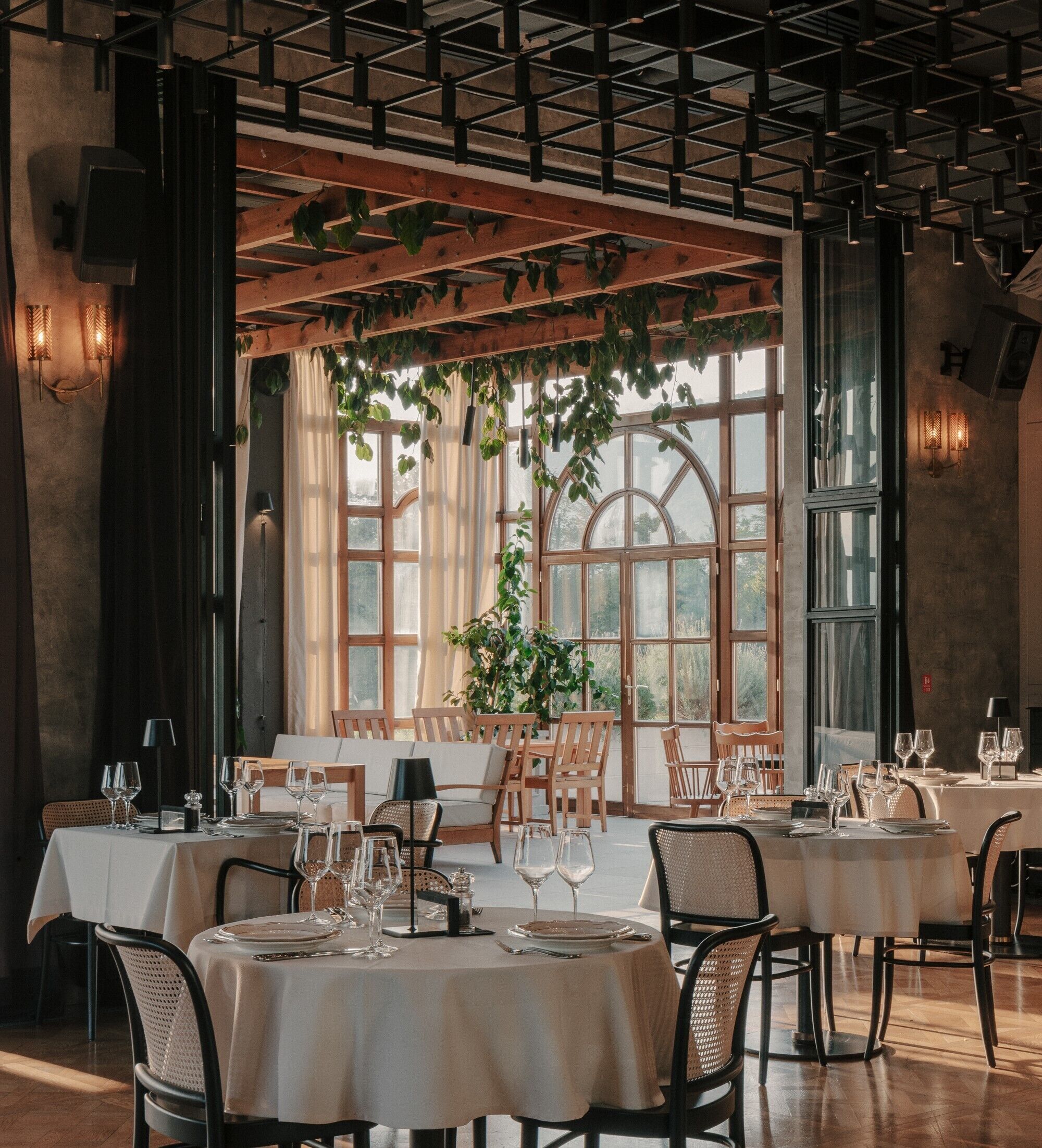A restaurant with tables set for dining, large windows, and hanging plants.