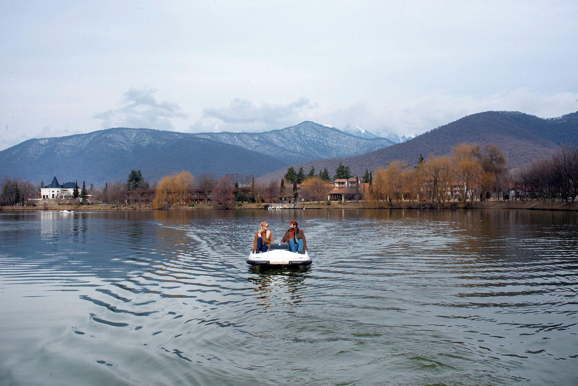 A lake with a boat carrying two people, surrounded by mountains and buildings.