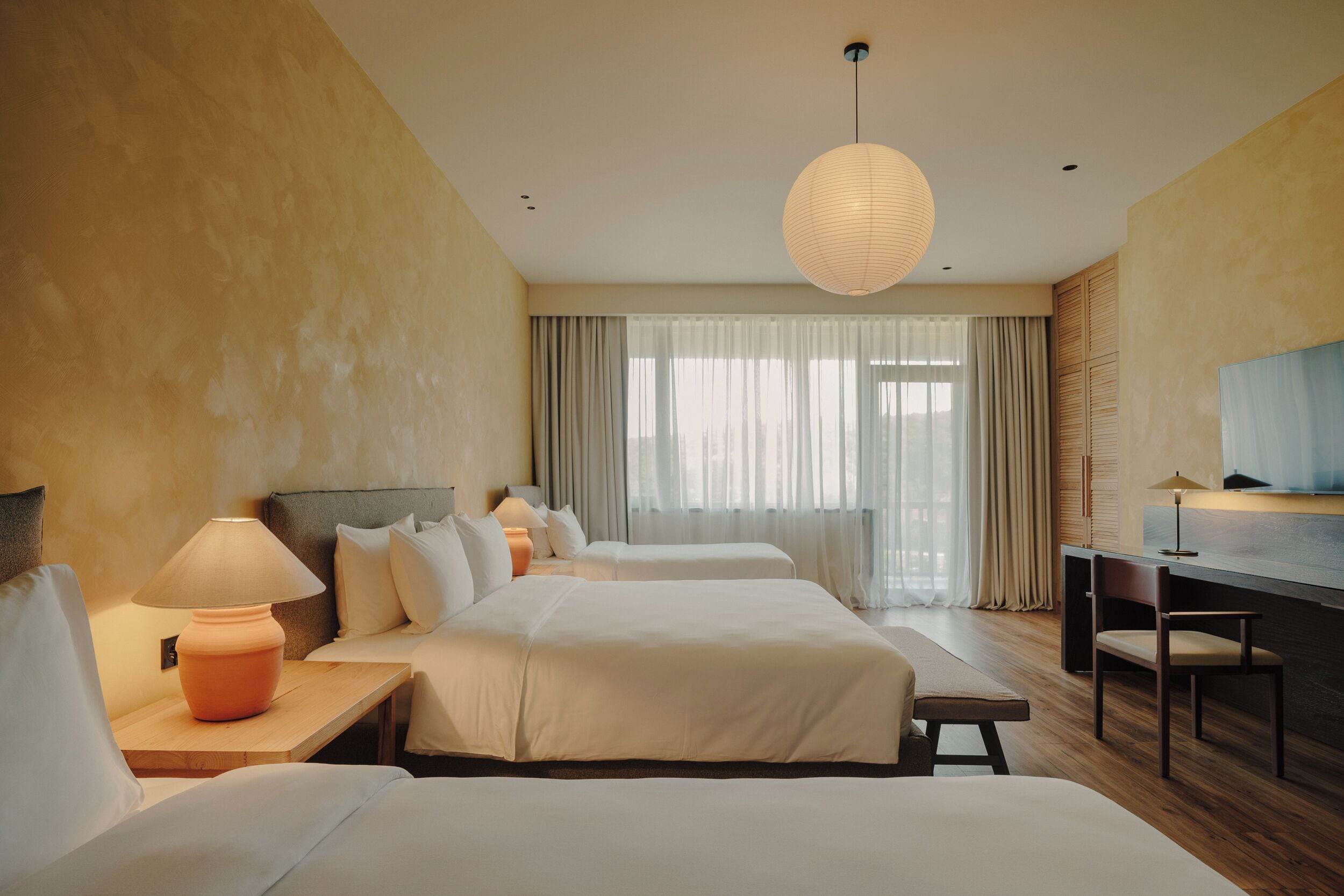 Ein Hotelzimmer mit zwei Betten, einem Schreibtisch, einem Stuhl, einer Lampe und einem großen Fenster mit Vorhängen.
