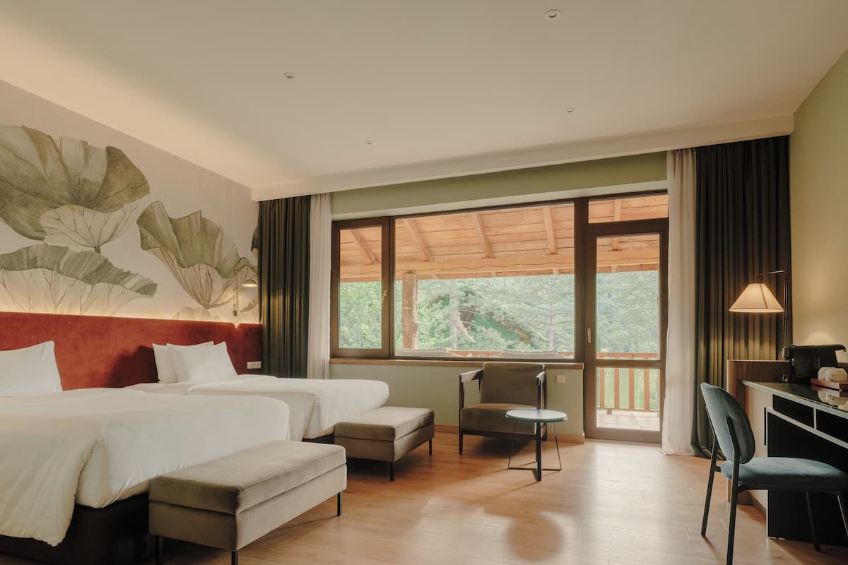 A hotel room with two beds, a desk, a chair, and a large window with a view of greenery.