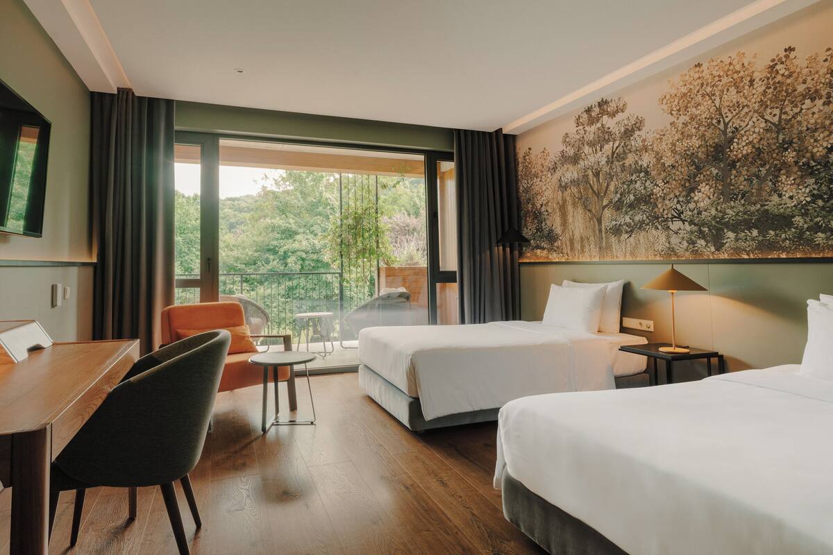 A hotel room with a balcony, a dining table, two beds, and a painting of trees.
