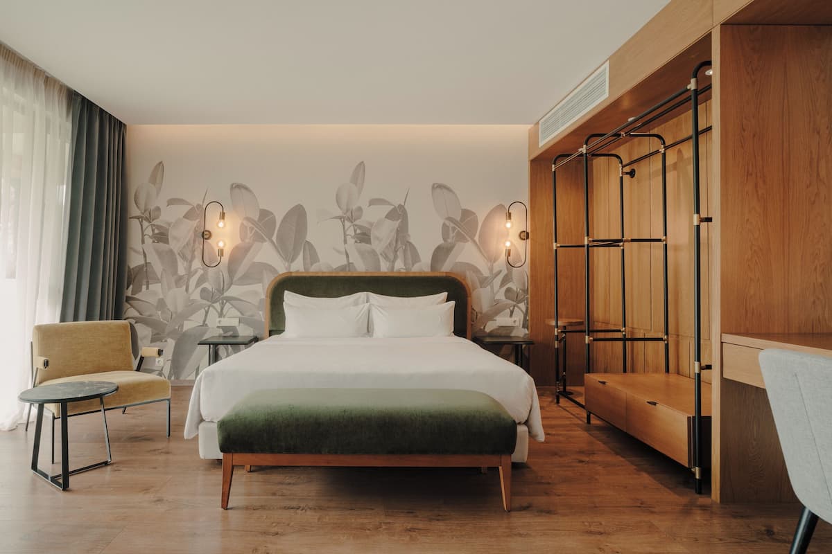 A hotel room with a bed, a chair, a nightstand, a wardrobe, and a wall with a leaf pattern.