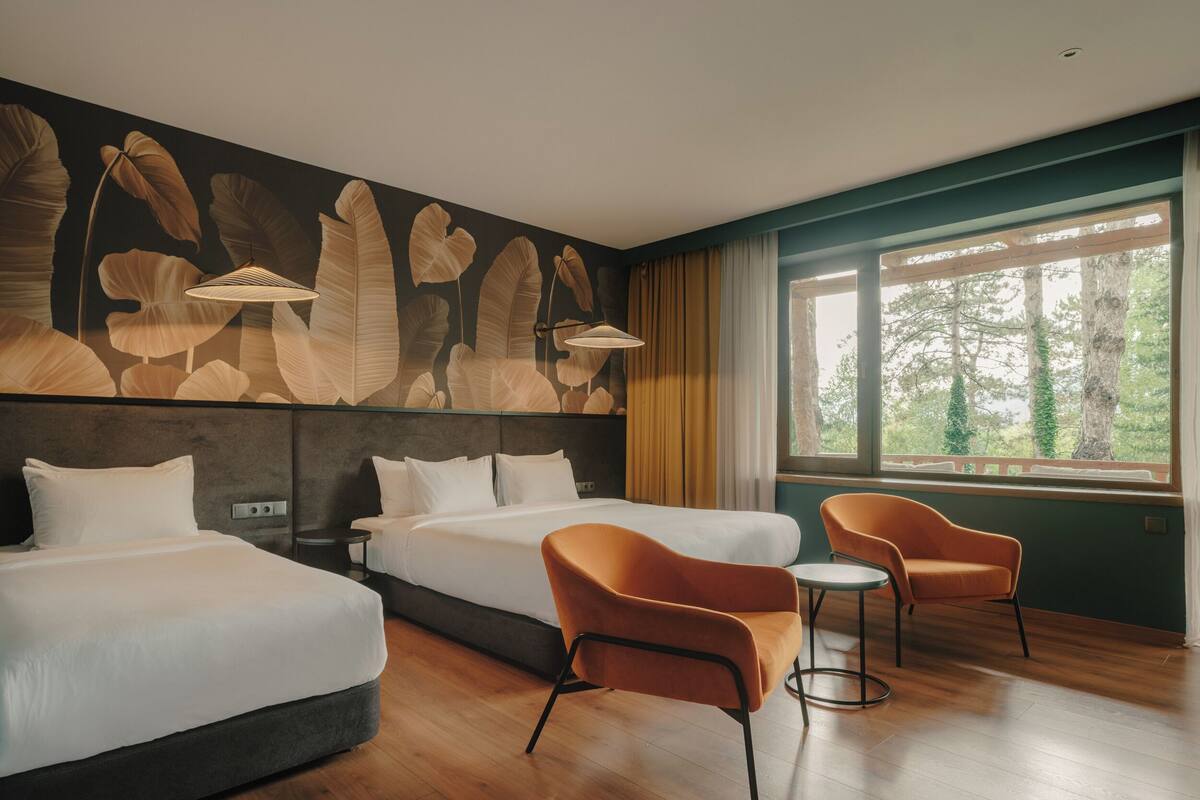 A hotel room with two beds, a small table, two chairs, a large window with a view of trees, and a wall with a leaf pattern.