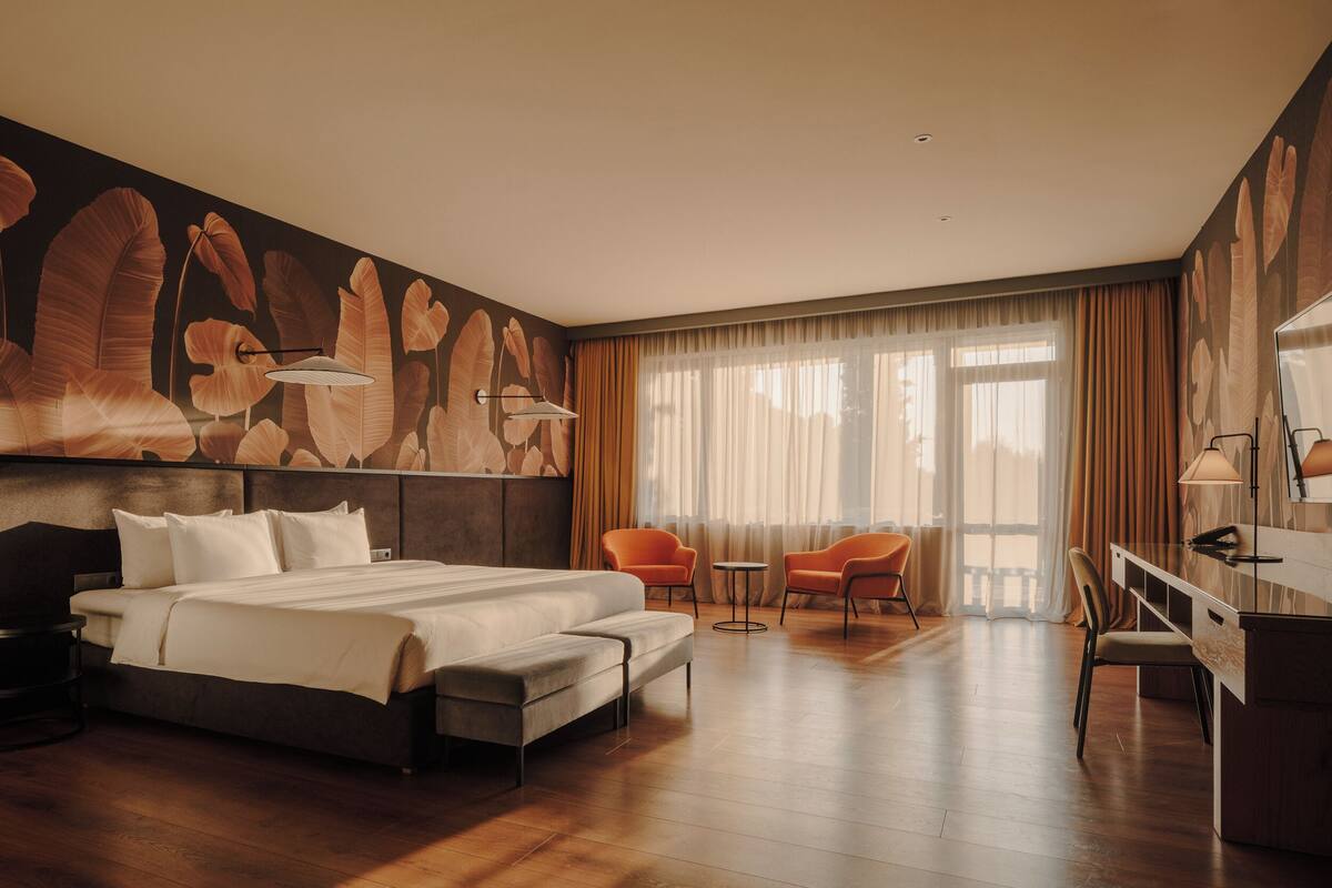 A hotel room with a large bed, two armchairs, a desk, and a large window with curtains.